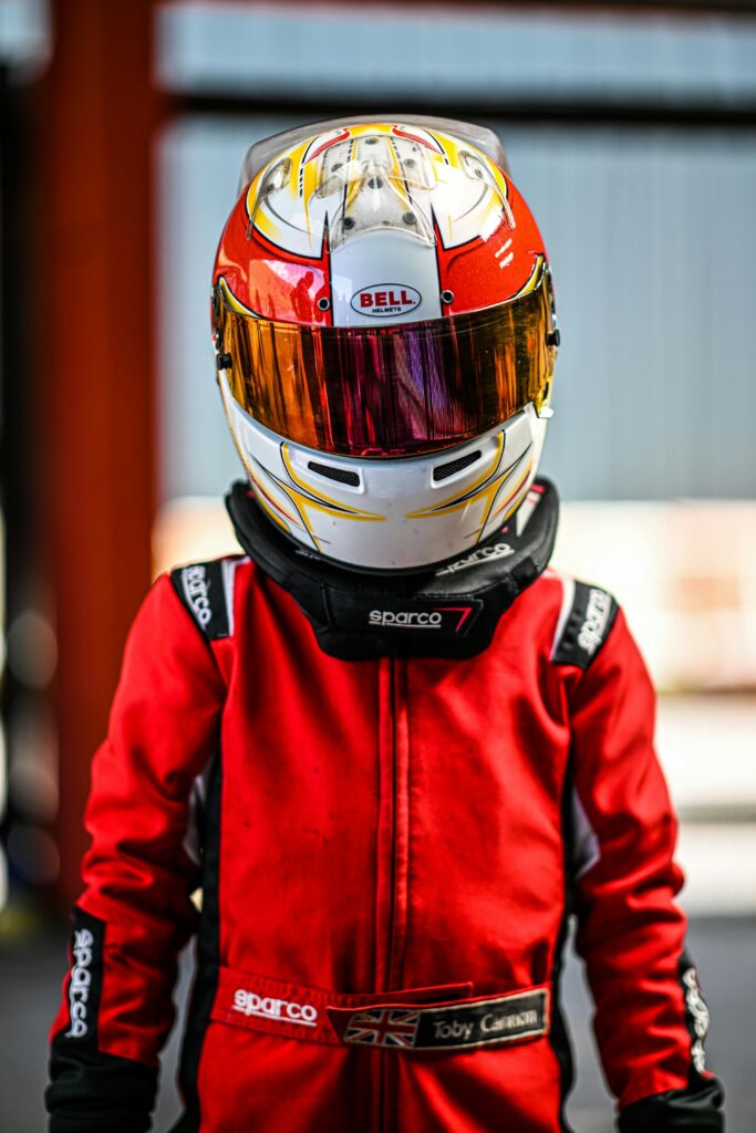 A child in a red racing suit and helmet stands ready for go-karting on a bright day.